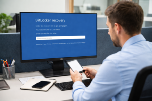 Office worker viewing a Windows BitLocker recovery screen on a desktop monitor while checking a phone for the recovery key